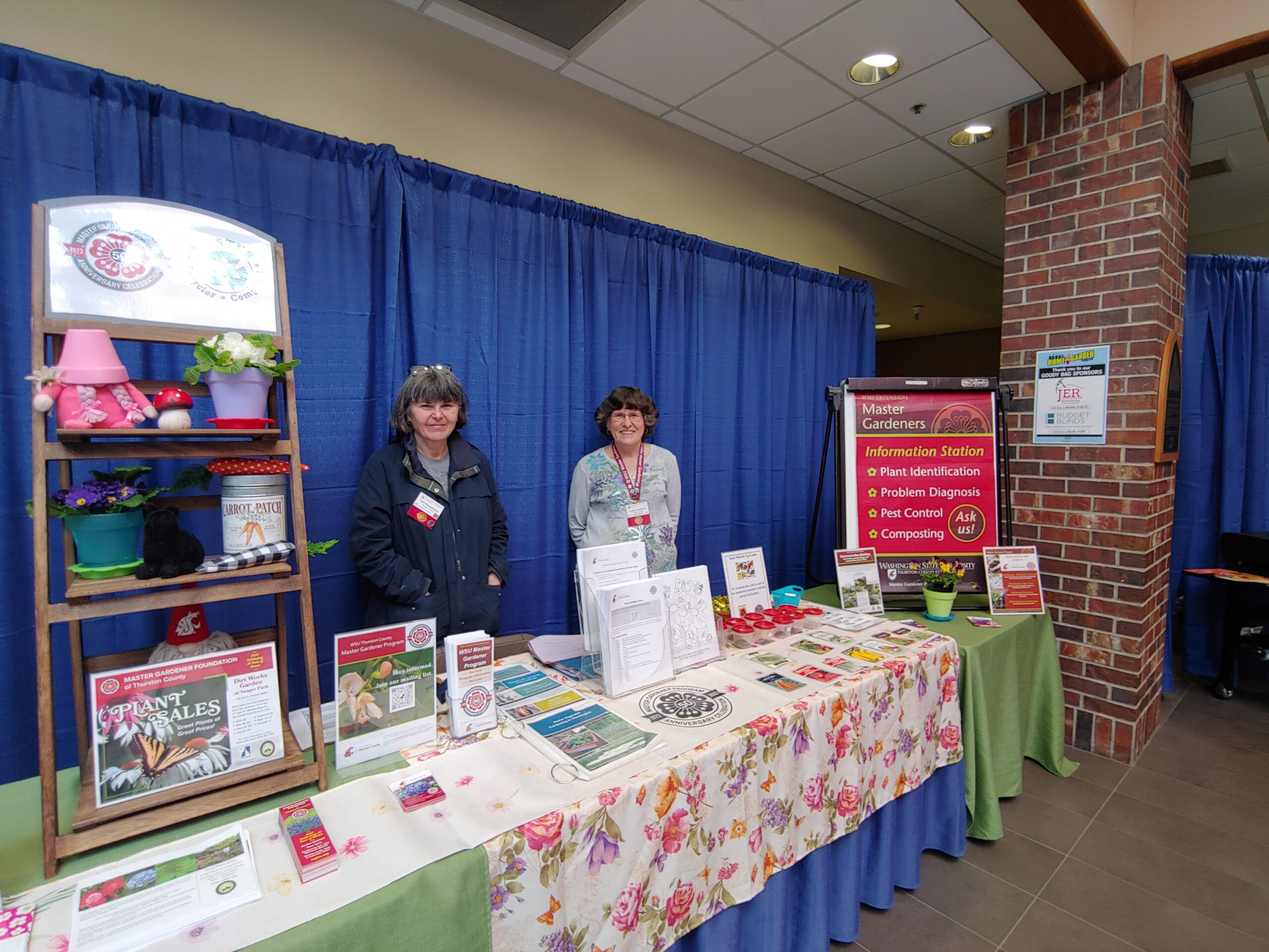 Two WSU Master Gardener volunteers smiling behind an information booth with gardening resources and decorative displays. 