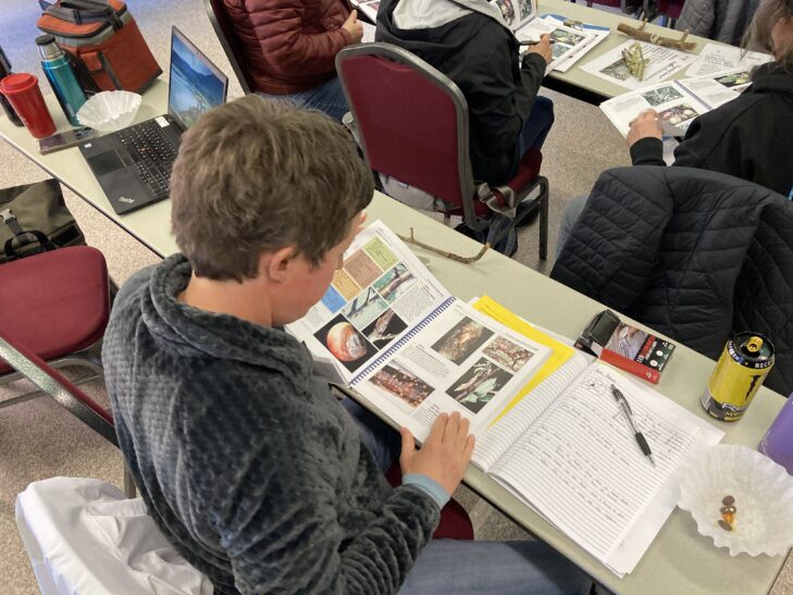 WSU Master Gardener volunteer looking at plant pathology book in a classroom, with notebook and pen on table. 