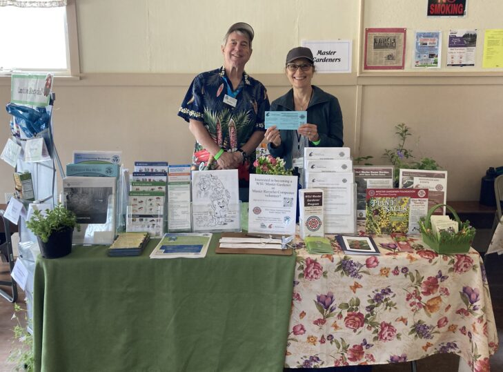 An WSU Master Gardener volunteer and a Master Recycler Composter volunteer smiling and standing behind a information table.