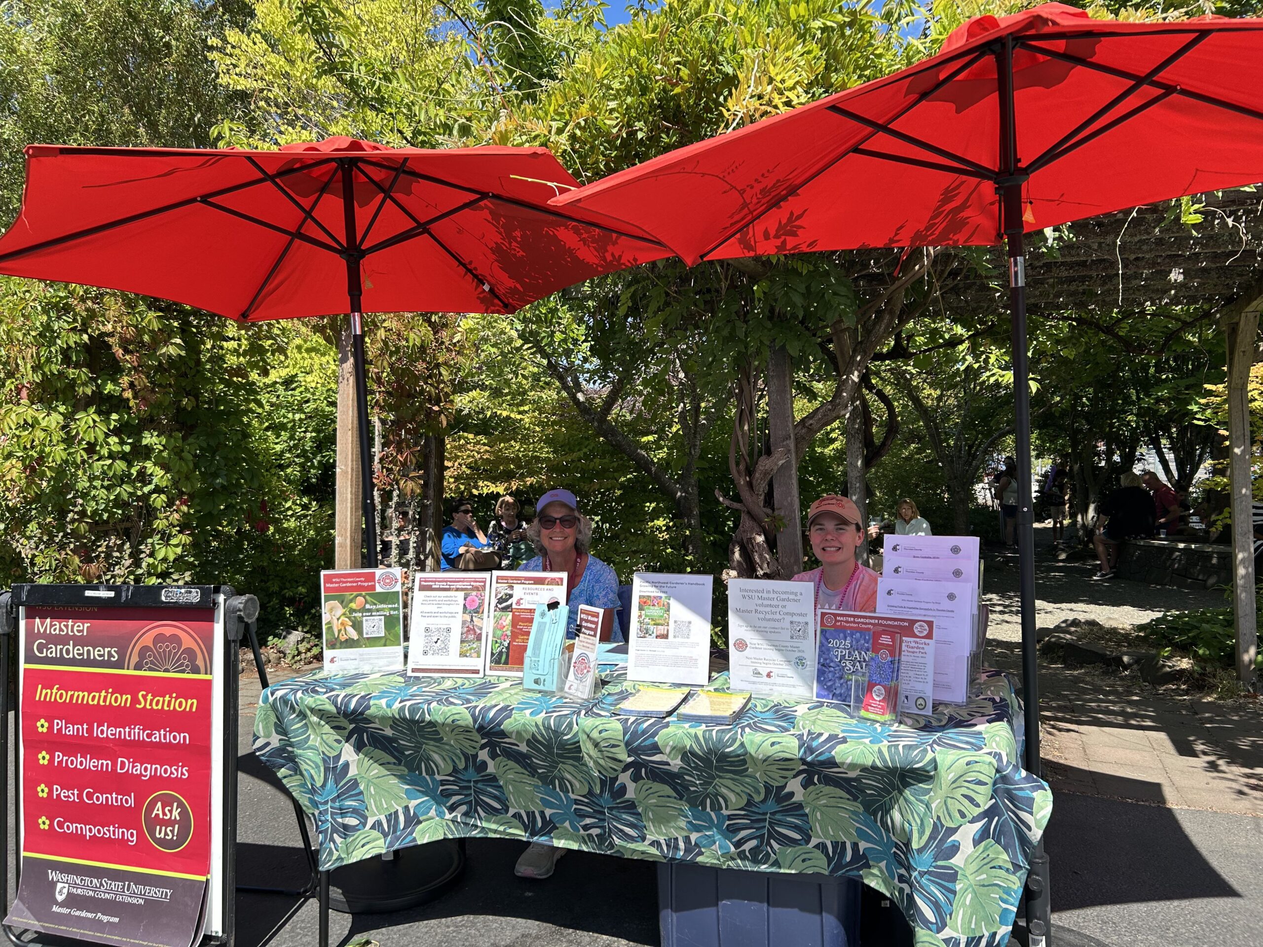 Two WSU Master Gardener volunteers smiling behind a table with gardening resources and event flyers at the Olympia Farmers Market.