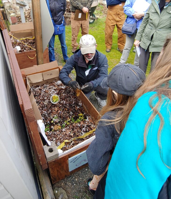 Master Recycler Composter volunteer showing workshop participants how to feed worms in a wooden worm bin.