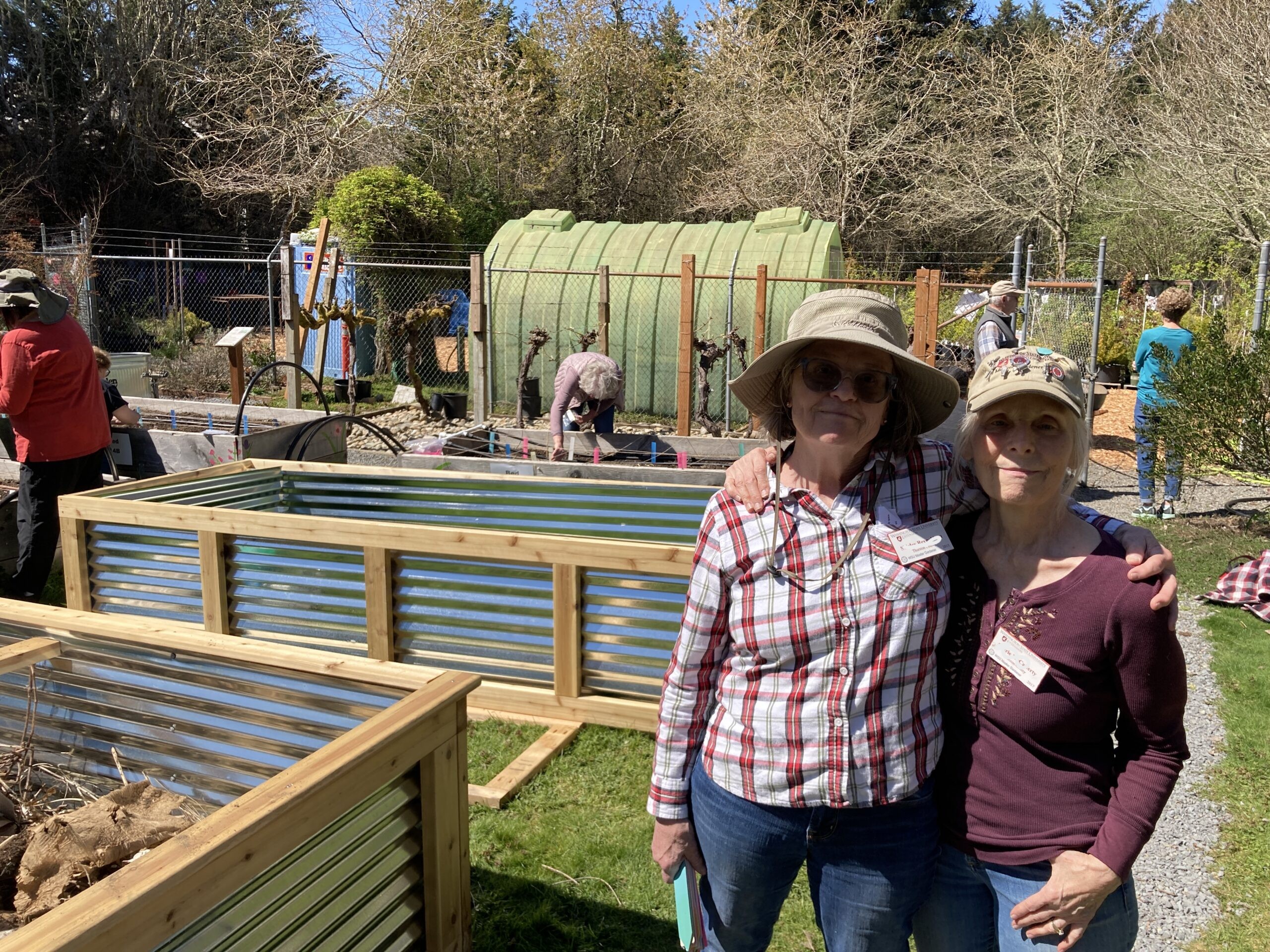 Two WSU Master Gardener volunteer smiling at the camera in front of newly constructed raised beds and older wooden raised beds with other volunteers planting.