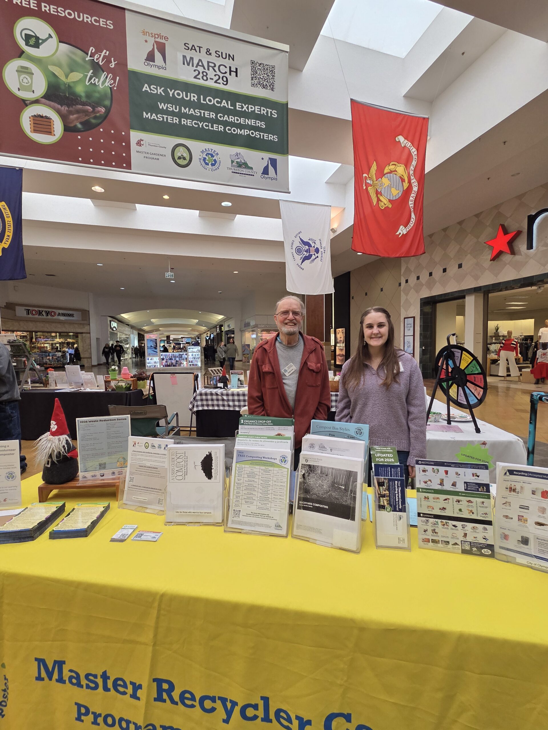 Two Master Recycler Composter volunteers smiling, standing behind a outreach booth with handouts on a yellow tablecloth.