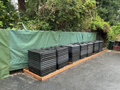 Compost bins lined up in front of canvas tarps hung on a chain link fence. 