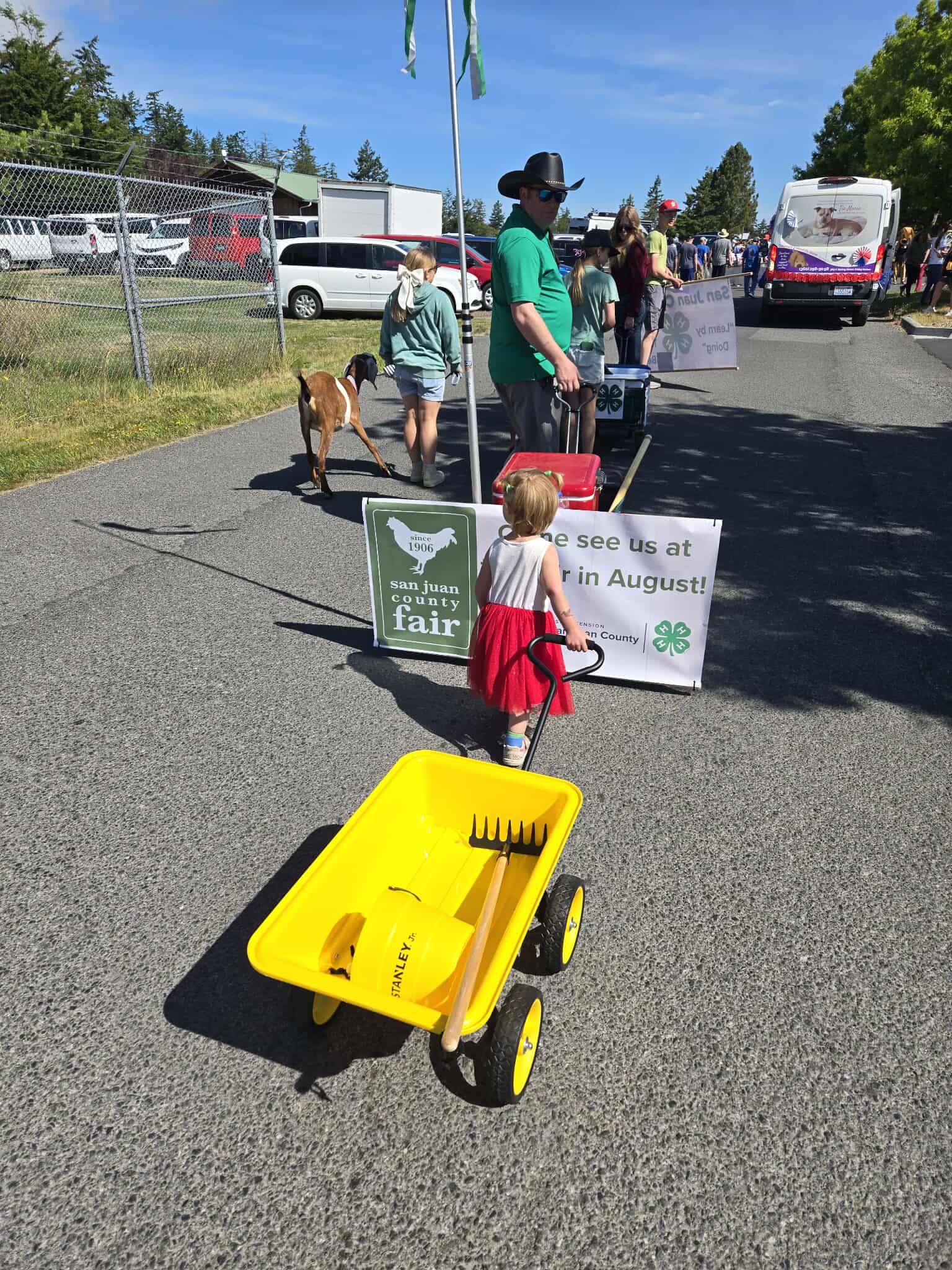 A small child pulls a yellow wagon with a rake, with adults, a goat, and fair signs in the background.
