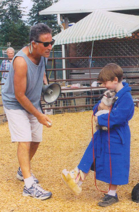 A man in sunglasses, a tank top, and shorts holds a megaphone and appears to be speaking to a child dressed in a blue robe and bunny slippers, holding a small rabbit. The scene is outdoors on wood chips, with a fence and a striped tent in the background.