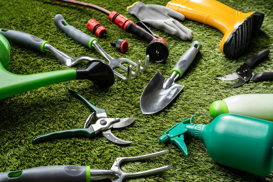 A high-angle close-up of various gardening and yard care tools lying on a surface of bright green artificial turf. The tools include a trowel, a cultivator, pruning shears, a watering can spout, hoses and nozzles, gloves, and a yellow rain boot.