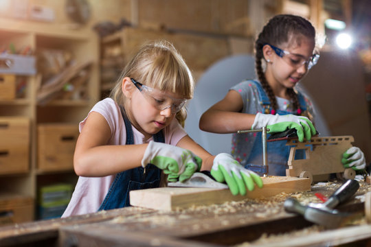 Two young girls in a woodworking shop, both wearing safety goggles and gloves. The girl in the foreground is using a hand planer on a wooden block, while the other girl works on a small, wooden toy truck.