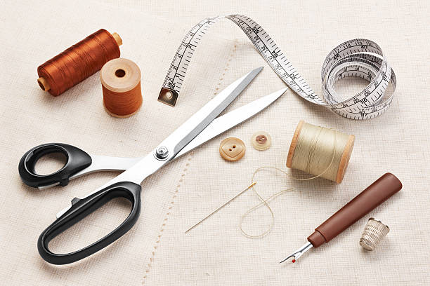 A flat lay photo of sewing tools on a light-colored canvas. The items include large fabric shears, a flexible measuring tape, spools of brown and tan thread, a needle, two wooden buttons, a thimble, and a seam ripper.