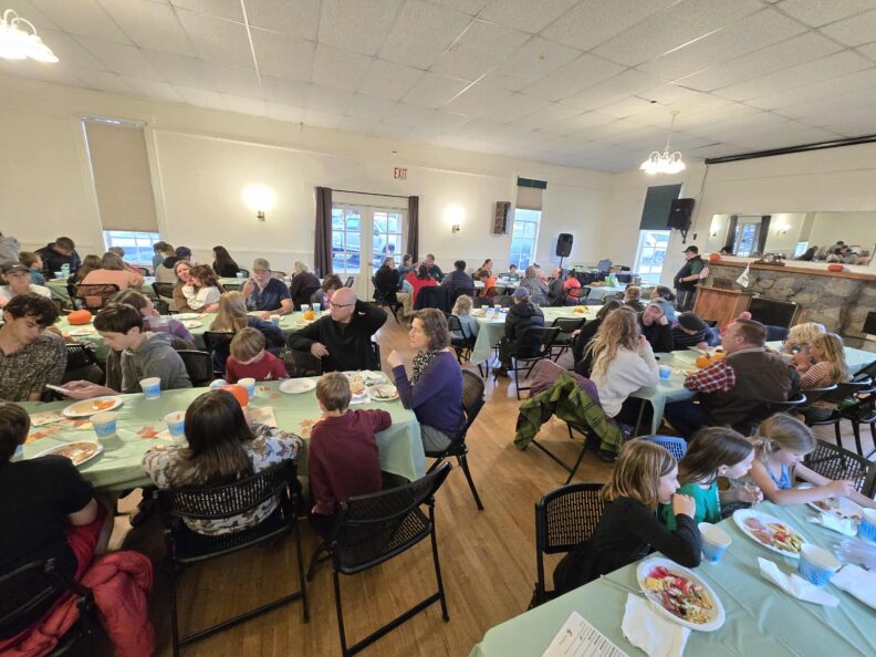 A wide-angle photo of a large group of adults and children sitting at long tables with pale green cloths, eating and socializing at an indoor community or banquet hall event.