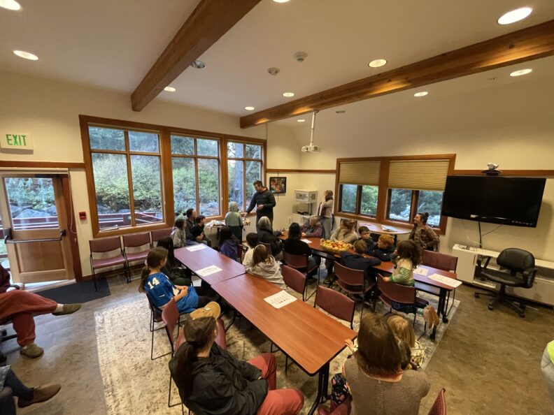 Wide-angle view of a group of adults and children sitting at tables in a brightly lit room with large windows, attentively watching an adult male standing and presenting near the center.