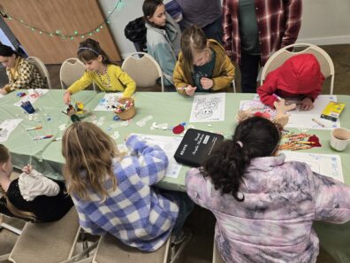 Children are seated at a long table covered with a light green tablecloth, busily coloring and crafting. The table is filled with various art supplies, including boxes of crayons, markers, and holiday-themed coloring pages. Adults are visible in the background, standing and watching the children work.