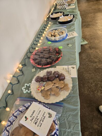 Several platters and containers of various cookies and treats are arranged on a long table covered with a light green tablecloth. The spread includes gingerbread men, chocolate-covered treats, brownies, and small round cookies, some labeled with "Allergy Info" cards. A string of warm white holiday lights is draped along the edge of the table.