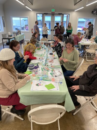 
A group of adults and children sit around long tables in a well-lit community room, engaged in a winter-themed craft activity. The tables are covered with light green cloths and scattered with craft supplies, including paper, stickers, and small decorations. Other people stand and talk in the background near a set of glass exit doors.