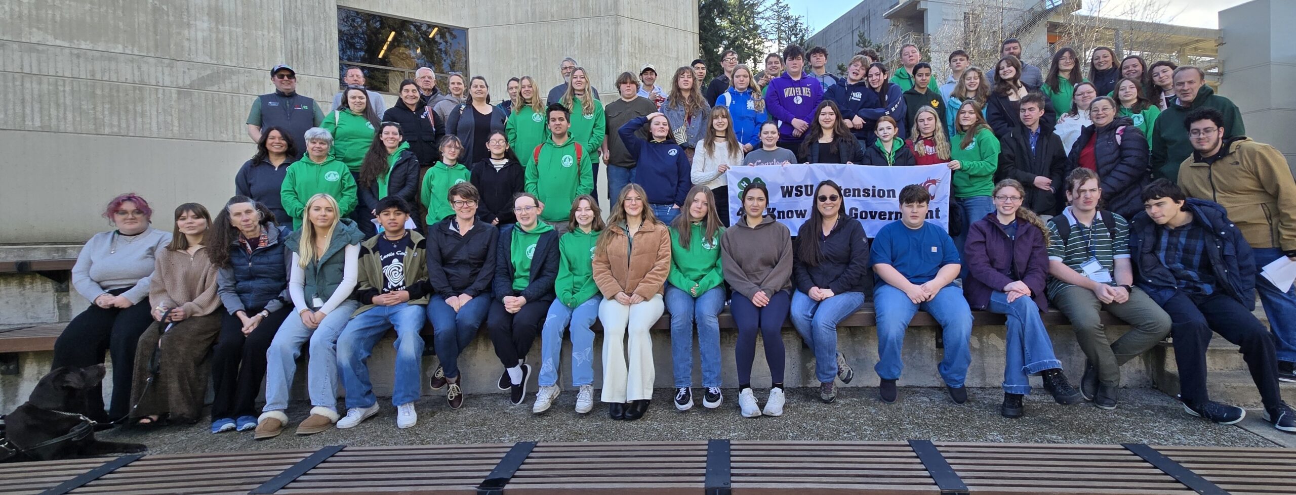 A wide group portrait of WSU Extension "Know Your Government" program participants posing on bleachers. The students and adults are wearing coordinated green and blue 4-H and WSU apparel while holding a program banner.