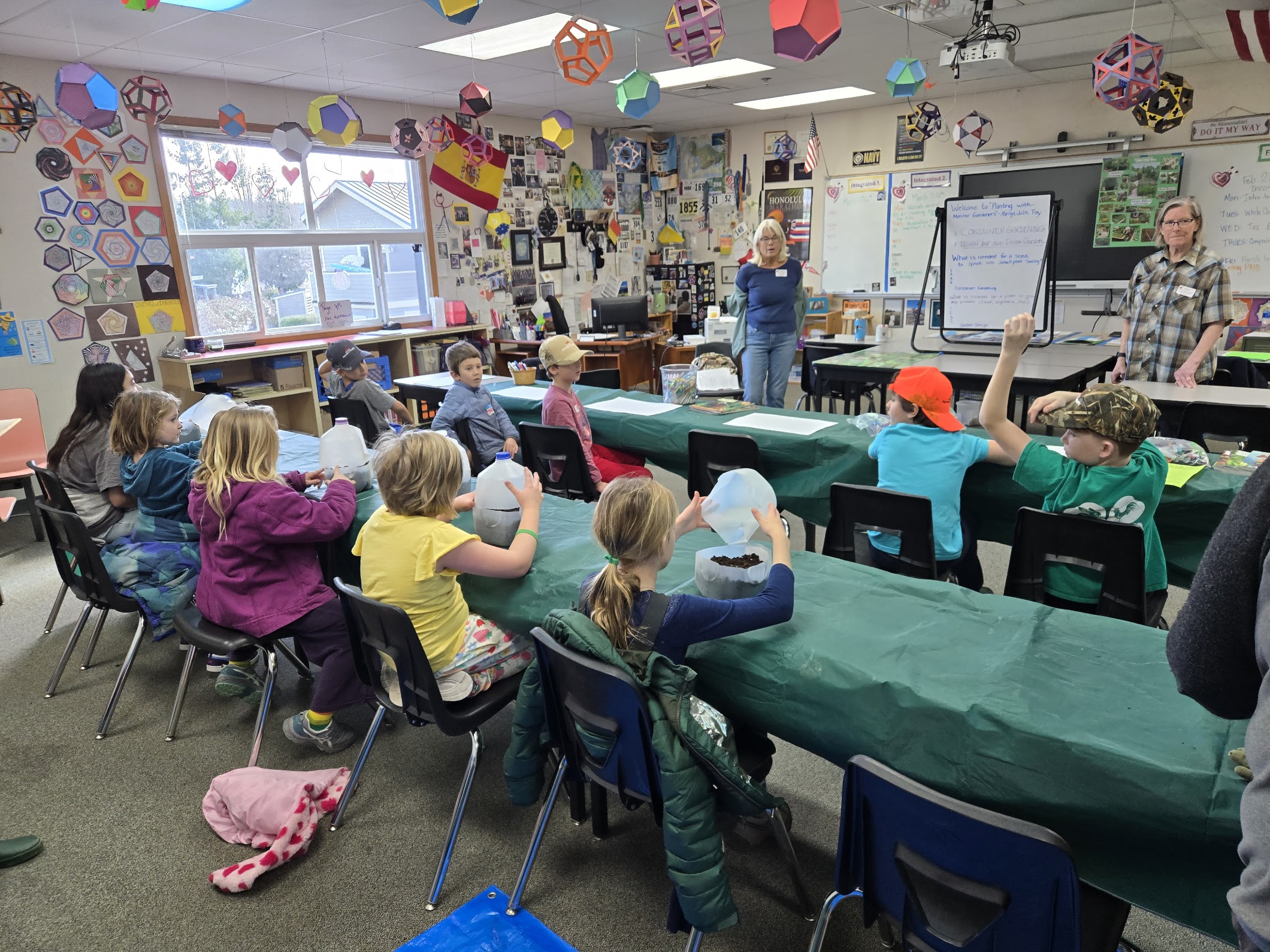 In a colorful classroom filled with student artwork and geometric ceiling decorations, two adult women stand at the front of the room leading a workshop. One woman stands near a whiteboard that reads "Welcome to Planting with Master Gardeners," while about ten children sit at long, green-clothed tables. The children are focused on the presentation, with some holding repurposed milk jugs and one student raising their hand to ask a question.