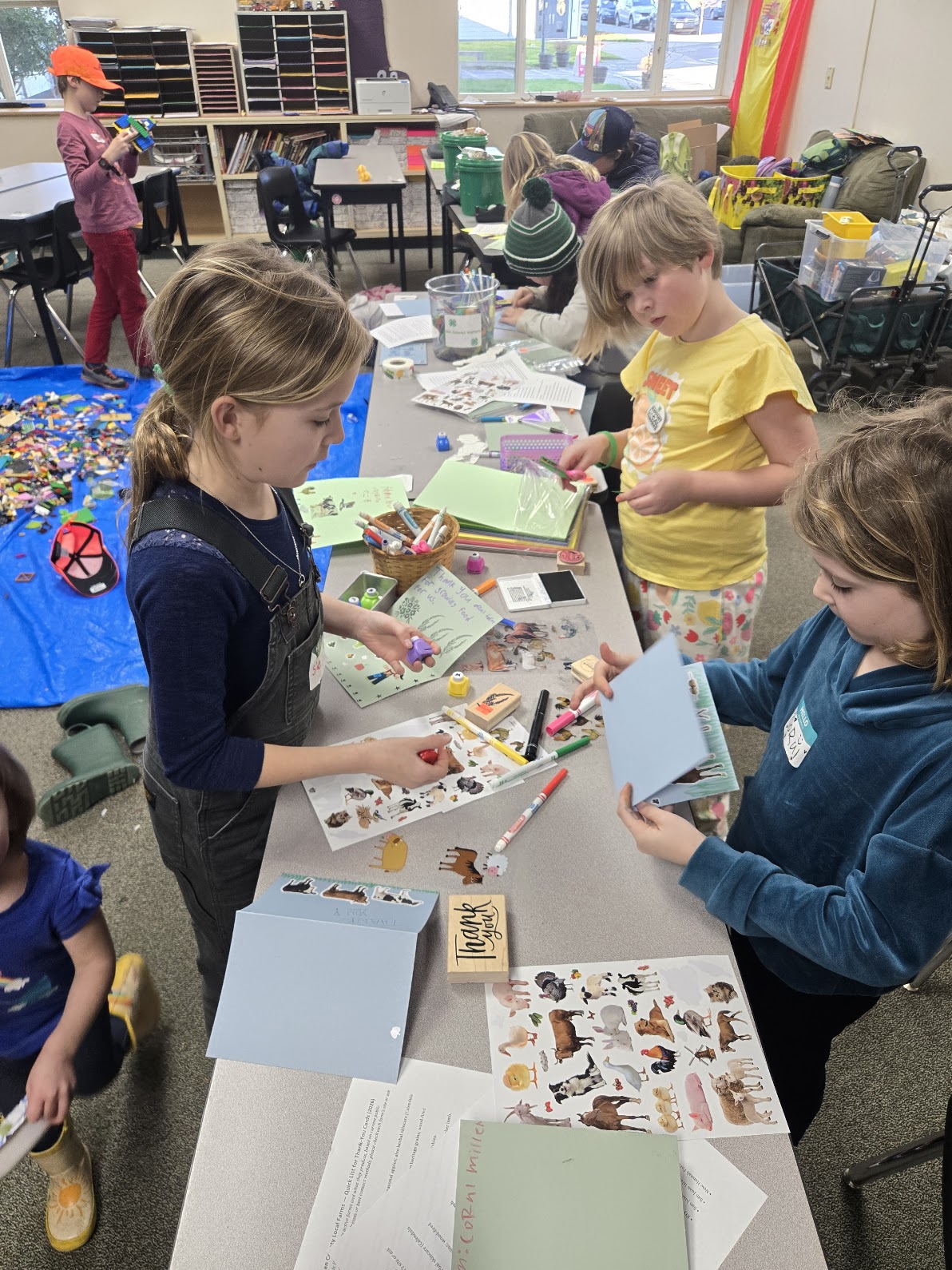 Three young girls are standing at a long table in a classroom, busy decorating thank-you cards. The table is covered in craft supplies, including sheets of animal stickers, rubber stamps, and markers. In the background, a blue tarp on the floor is covered in LEGO bricks for the decompression activity.