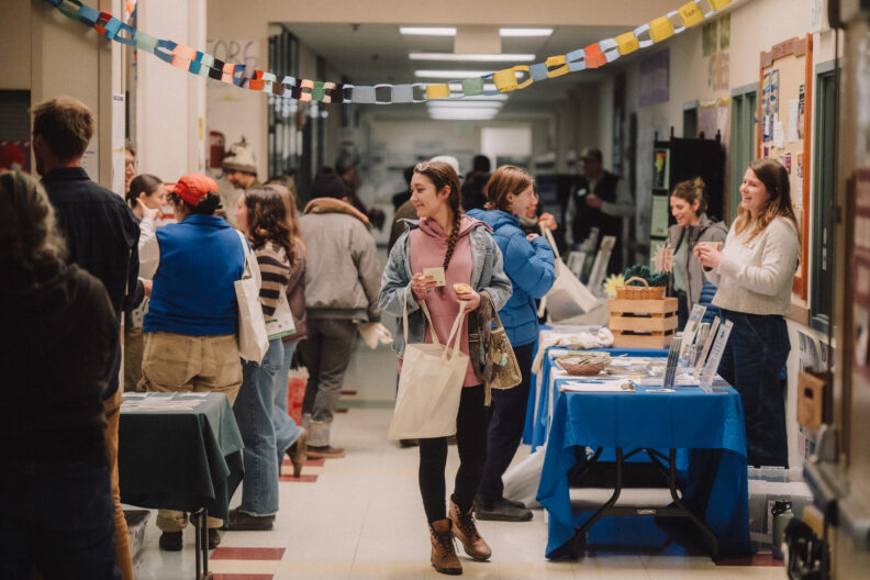 Image of a woman walking through the trade show at the 2026 SJI Ag Summit. There are info tables and service providers on either side of the hallway of the Friday Harbor High School