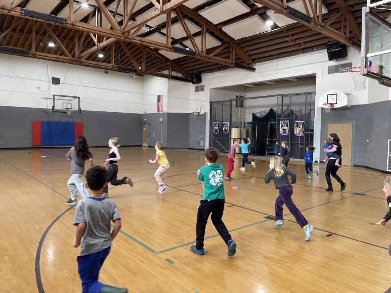 A group of children and several adults are active in a gymnasium with a high, wood-truss ceiling. Several of the children are running across the polished wooden floor, including one child in the foreground wearing a green t-shirt with a white 4-H clover logo on the back. A basketball hoop and a large American flag are visible in the background.