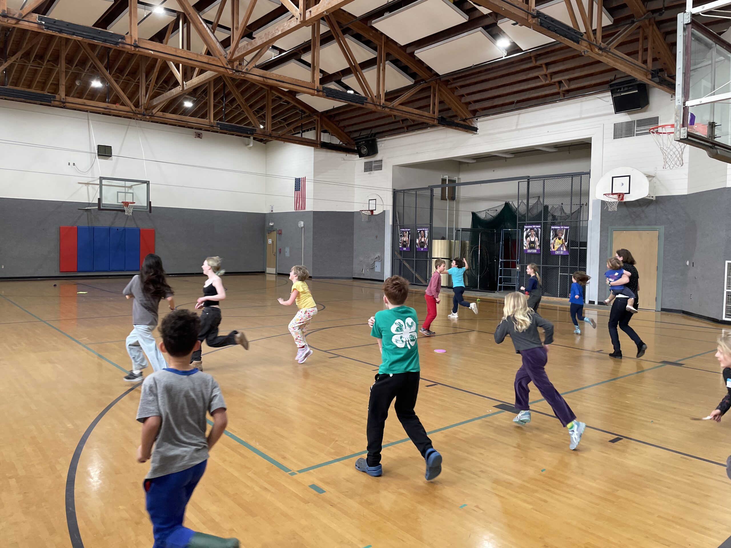 A group of children and several adults are active in a gymnasium with a high, wood-truss ceiling. Several of the children are running across the polished wooden floor, including one child in the foreground wearing a green t-shirt with a white 4-H clover logo on the back. A basketball hoop and a large American flag are visible in the background.
