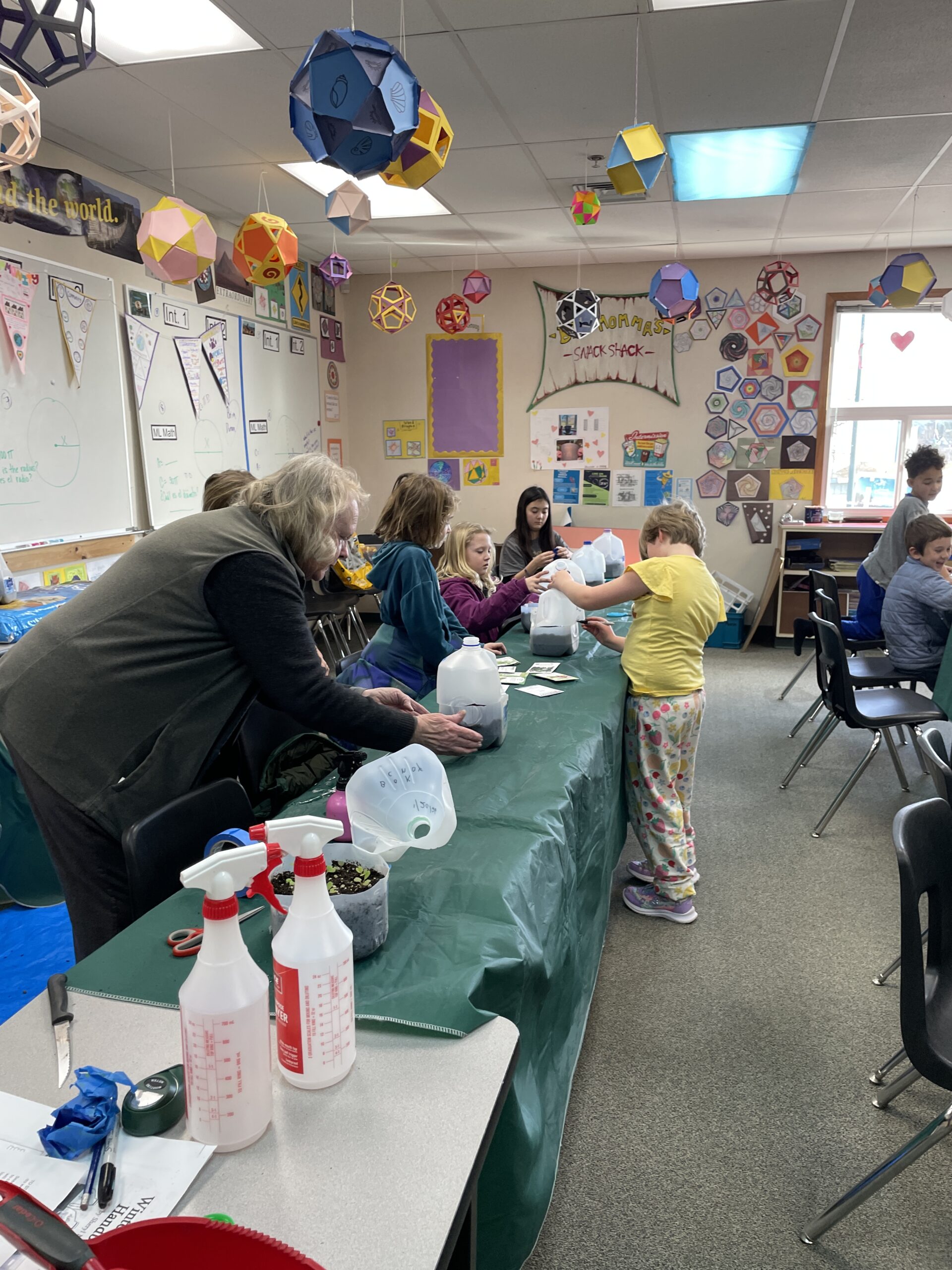 Inside a classroom decorated with colorful geometric paper lanterns hanging from the ceiling, several children and an adult woman are working at a long table covered in a green cloth. They are focused on a "seed starting" project using recycled plastic milk jugs filled with soil. Spray bottles and seed packets are visible on the table.