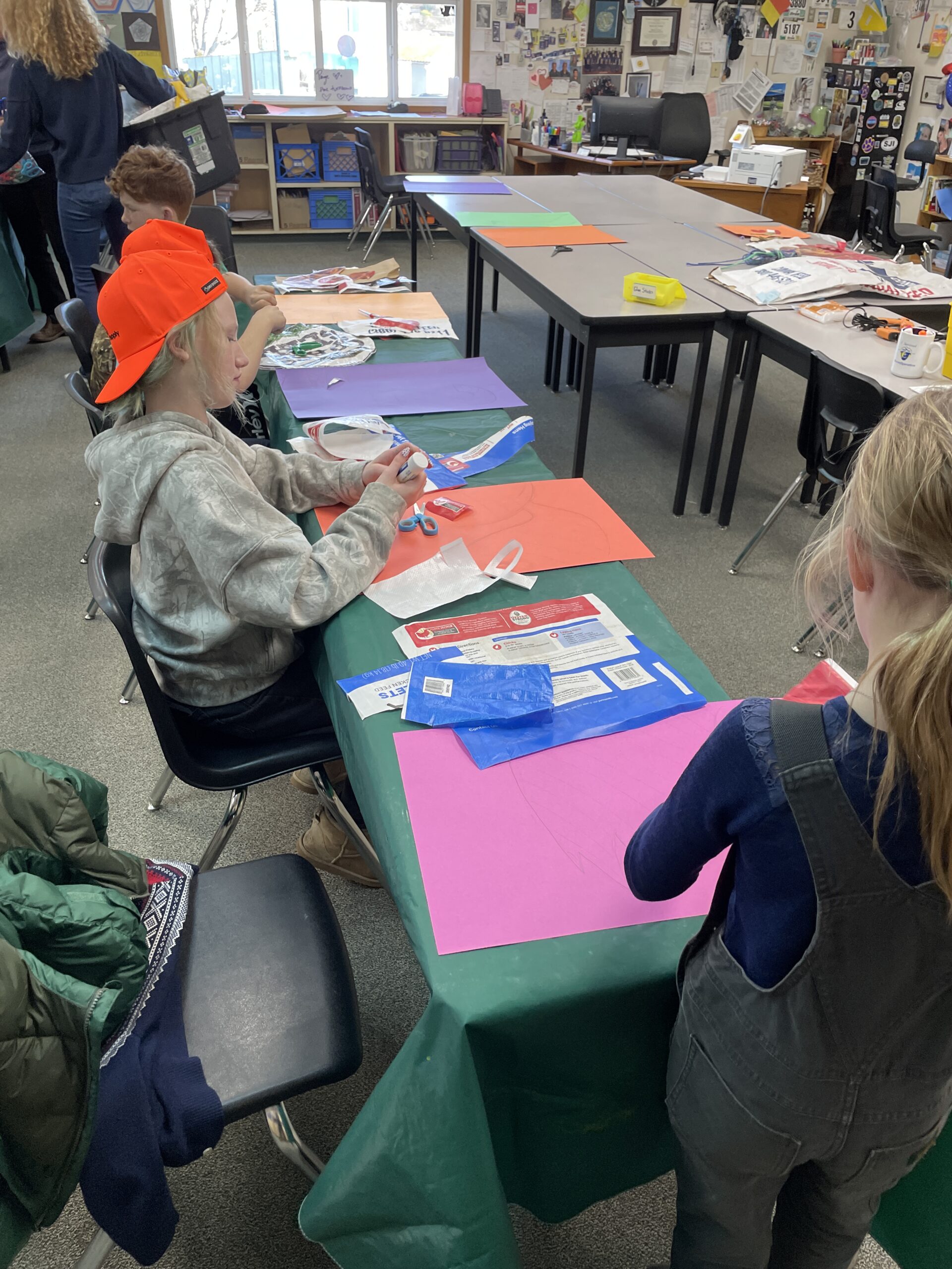 In a brightly lit classroom, two children sit at a table covered with a green cloth, working on an art project. One child, wearing an orange hat and a grey hoodie, is focused on gluing materials onto a large piece of orange paper. On the table are various recycled materials, including colorful blue and red plastic feed bags, scraps of paper, and scissors. In the background, other tables and classroom supplies are visible.