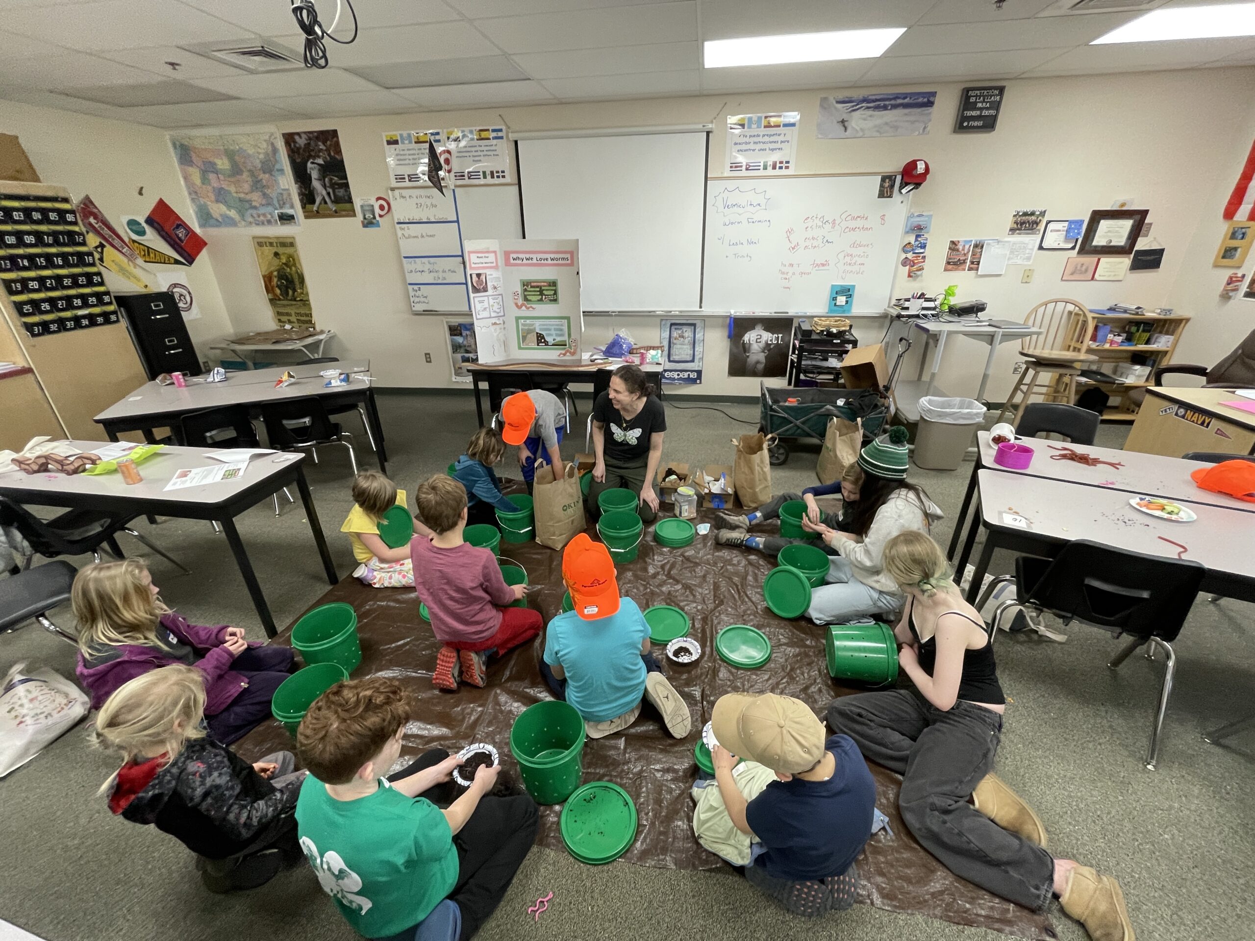 In a classroom, an adult instructor and about a dozen children are sitting in a circle on a large brown tarp spread across the floor. Each child has a bright green plastic bucket and is focused on assembling an in-ground worm bin. A tri-fold poster board with the title "Why We Love Worms" stands on a desk in the background, alongside a whiteboard with notes on vermiculture.