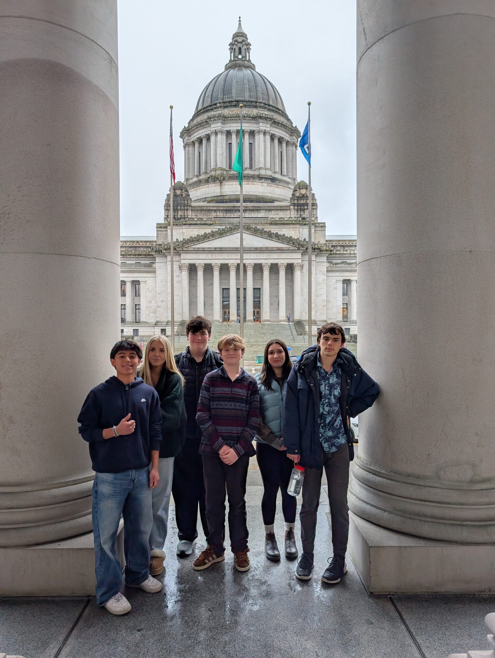 A group of six teenagers stands between two large stone pillars, posing in front of the Washington State Legislative Building in Olympia. The domed capitol building and three flags are visible in the background under an overcast sky.