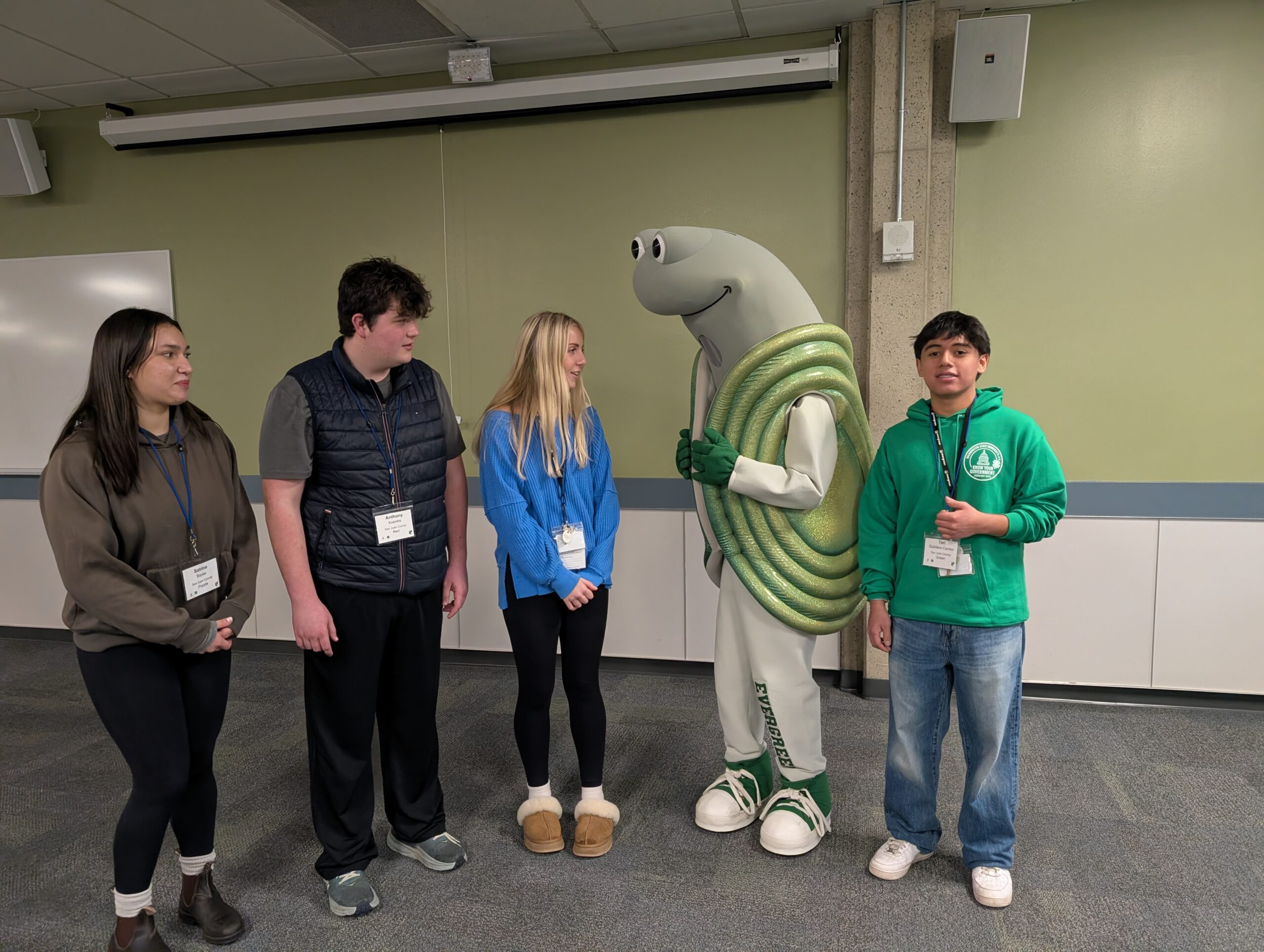 Four teenagers stand in a room with "Speedy the Geoduck," the mascot of The Evergreen State College. The students are wearing conference lanyards and casual clothes, while the mascot is a large, grey, smiling geoduck with a green coiled shell and white track suit.