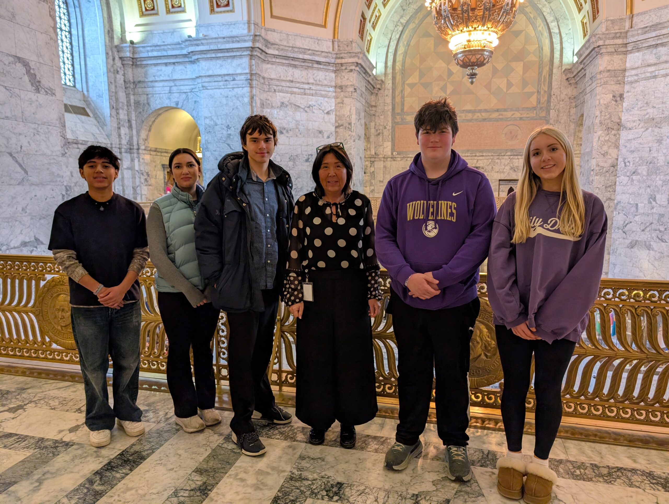 State Representative Debra Lekanoff stands with five San Juan County 4-H teenagers on a marble balcony inside the Washington State Legislative Building. Representative Lekanoff wears a black and white polka-dot blouse. They are posed in front of an ornate gold railing with a large, glowing chandelier and decorative arched architecture in the background.