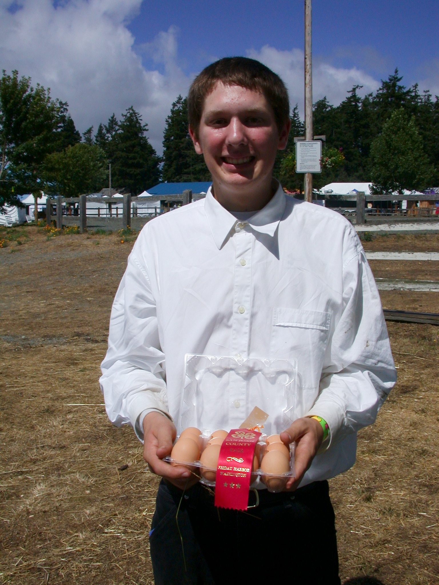 A smiling young man in a white dress shirt holding a carton of brown eggs decorated with a red award ribbon at an outdoor county fair.