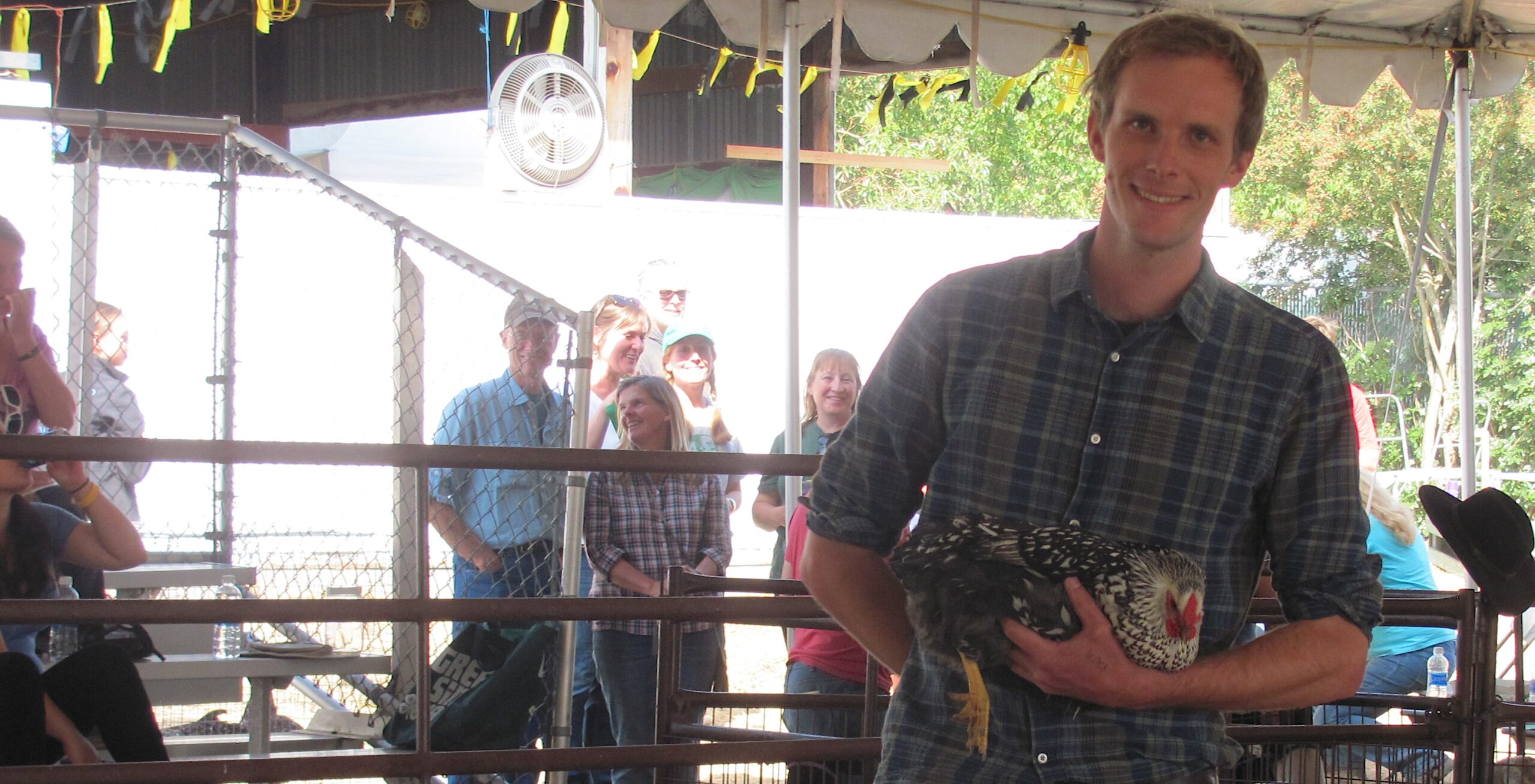 A smiling man in a plaid shirt holding a speckled silver and black chicken under a tent at an outdoor event.