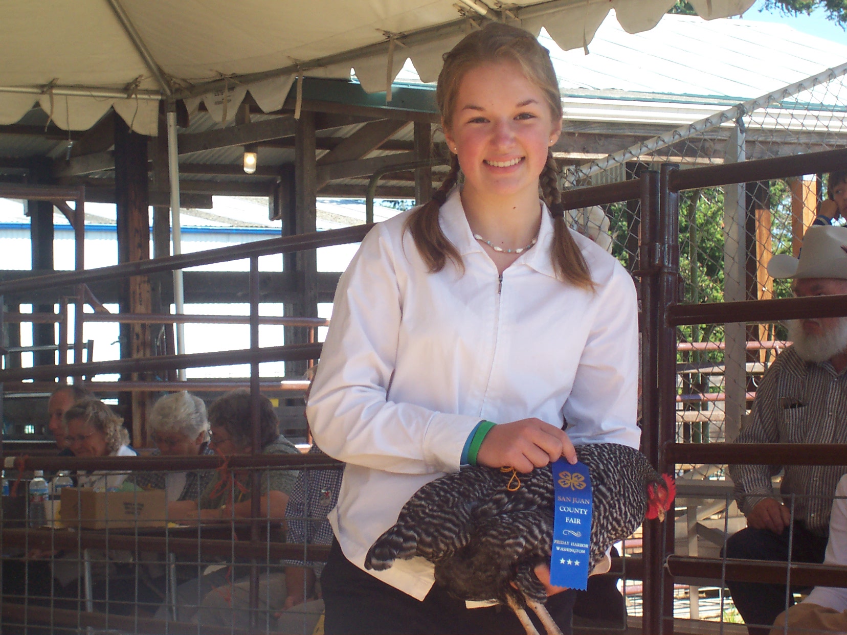 A smiling teenage girl with braided hair and a white jacket holds a Barred Rock chicken displaying a blue first-place ribbon from the San Juan County Fair.