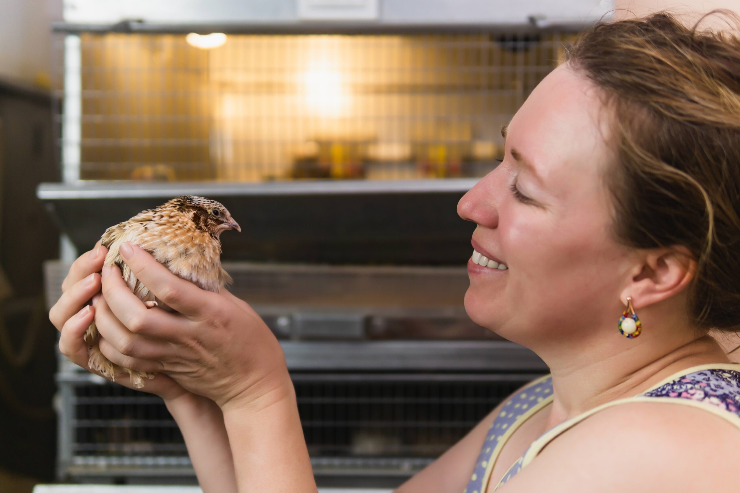 A smiling woman gently holding a small, brown-speckled quail in her hands with metal bird cages in the background.