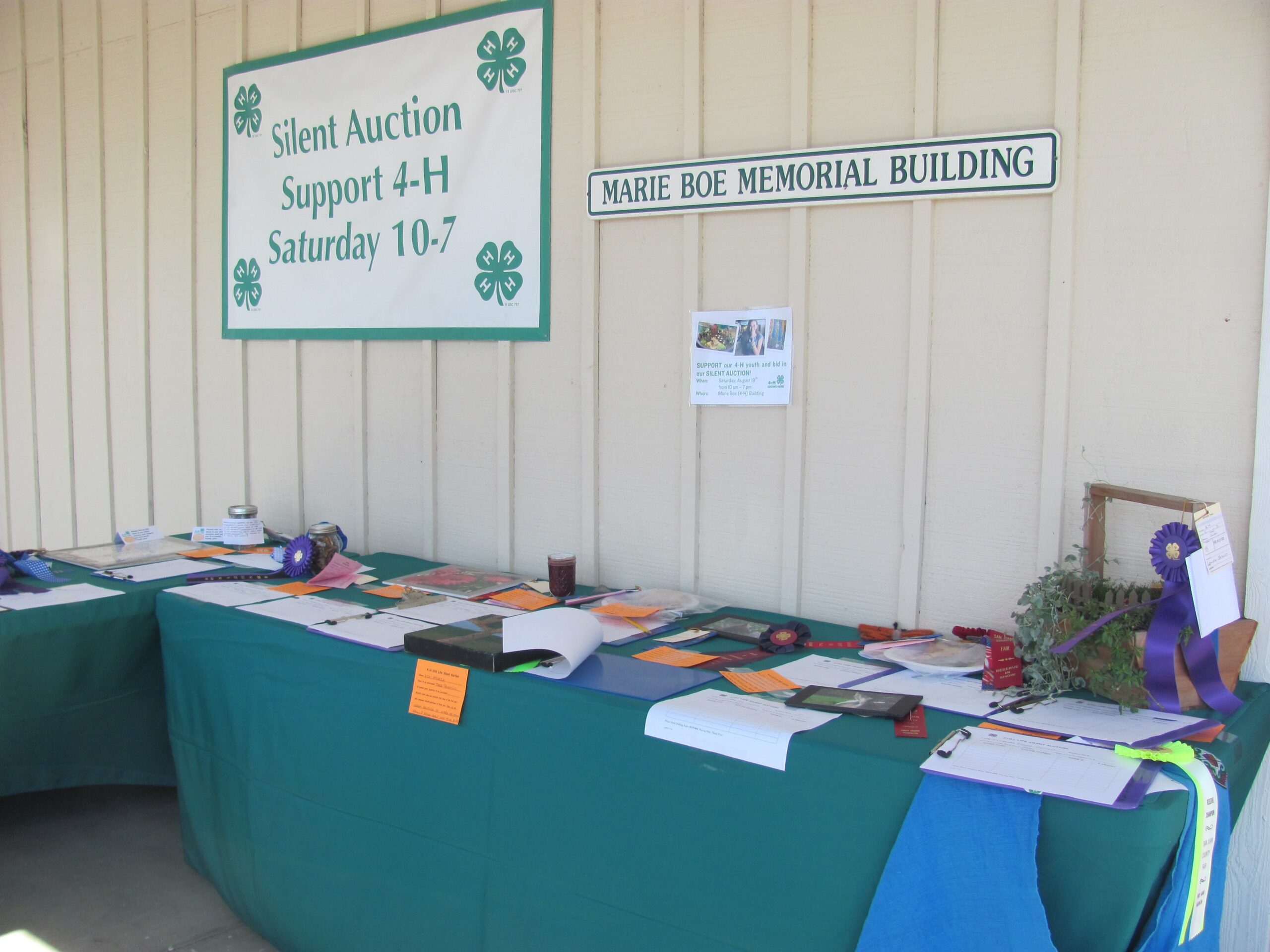 Gemini said A long table with a green tablecloth holds various silent auction items, bid sheets, and ribbons inside the Marie Boe Memorial Building, with a large sign overhead reading "Silent Auction Support 4-H Saturday 10-7."