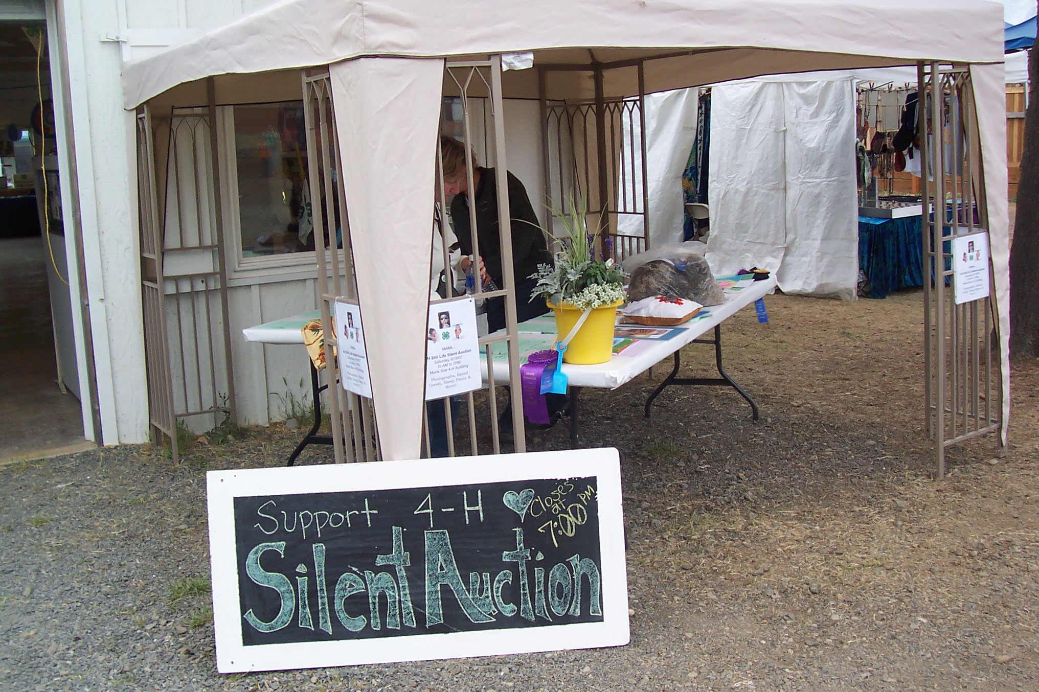 A chalkboard sign in the foreground reads "Support 4-H Silent Auction" with a 7:00 PM closing time, positioned in front of a white tent where auction items are displayed on a folding table.