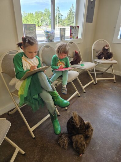 wo young girls sit on folding chairs under a window, writing on clipboards with pencils. A stuffed animal sits on a nearby chair, and a realistic fake porcupine prop rests on the floor in front of them.