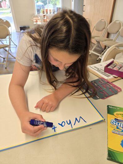 Close-up of a young girl leaning over a table, using a blue marker to write "What" on a large white poster board, with markers and stencils nearby.