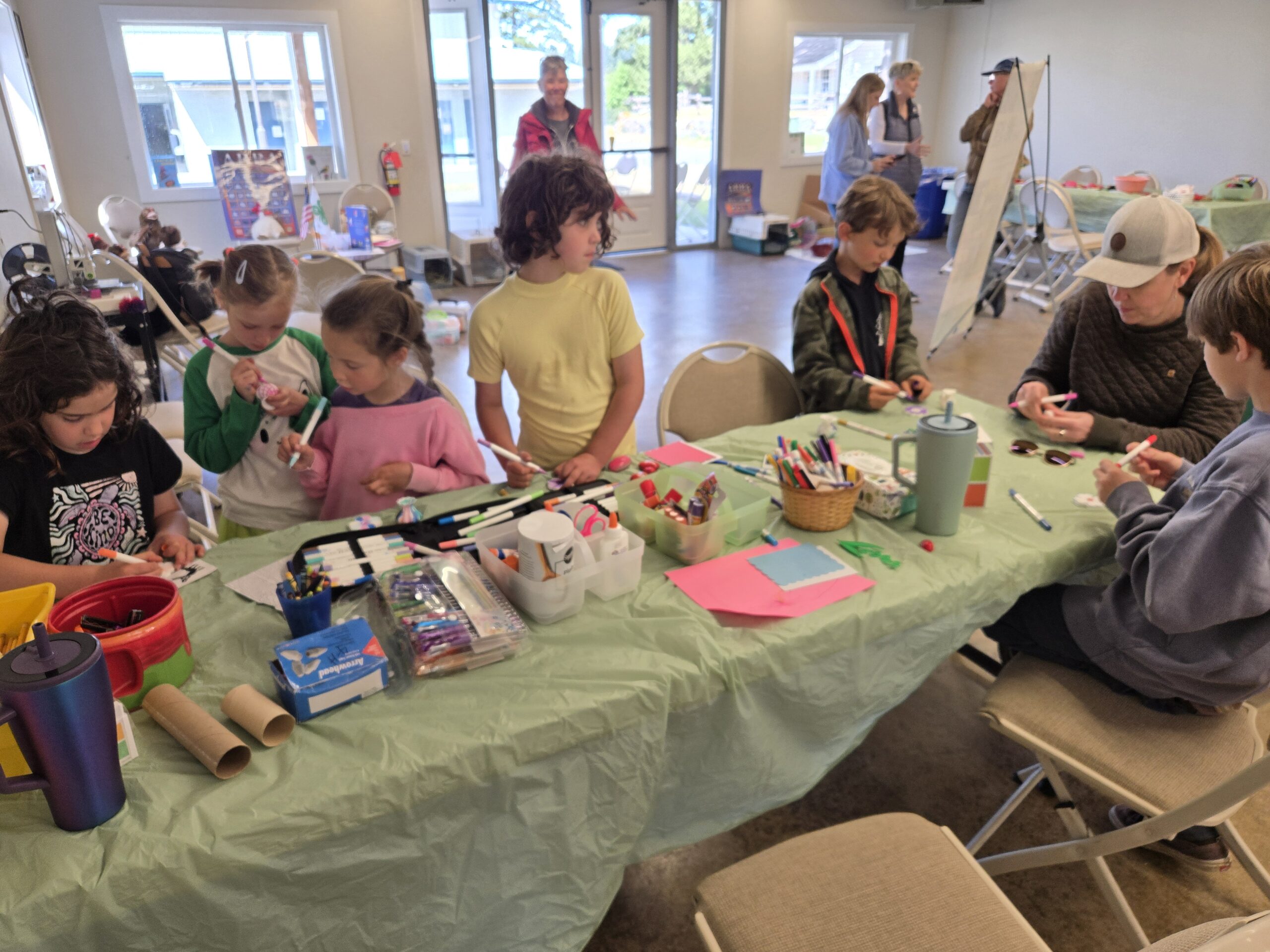 A group of children and an adult sit and stand around a long table covered in a light green tablecloth, engaged in an arts and crafts activity with markers, paper, and various craft supplies.