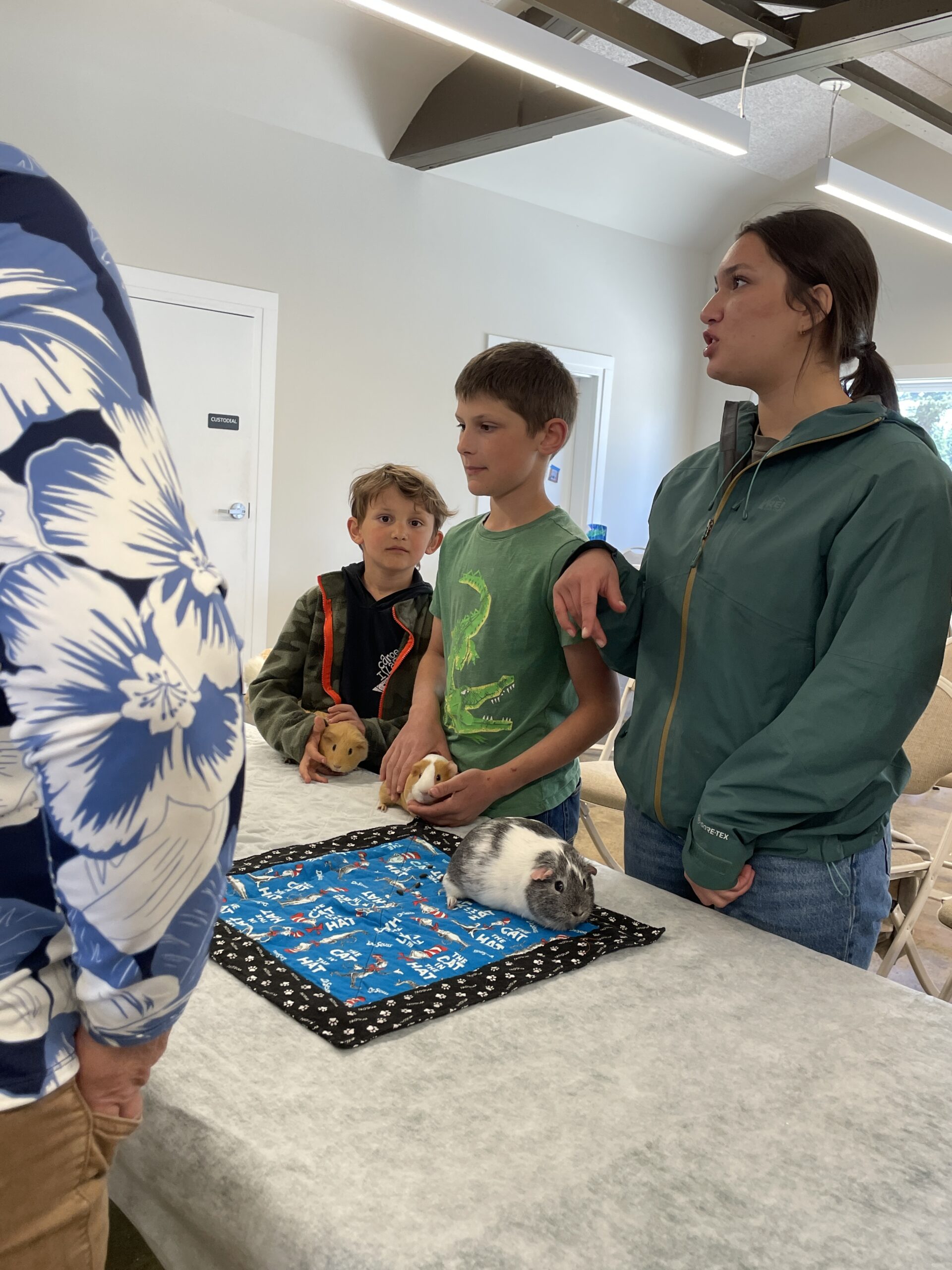 Two young boys and a woman standing at a table, interacting with guinea pigs during an educational presentation.