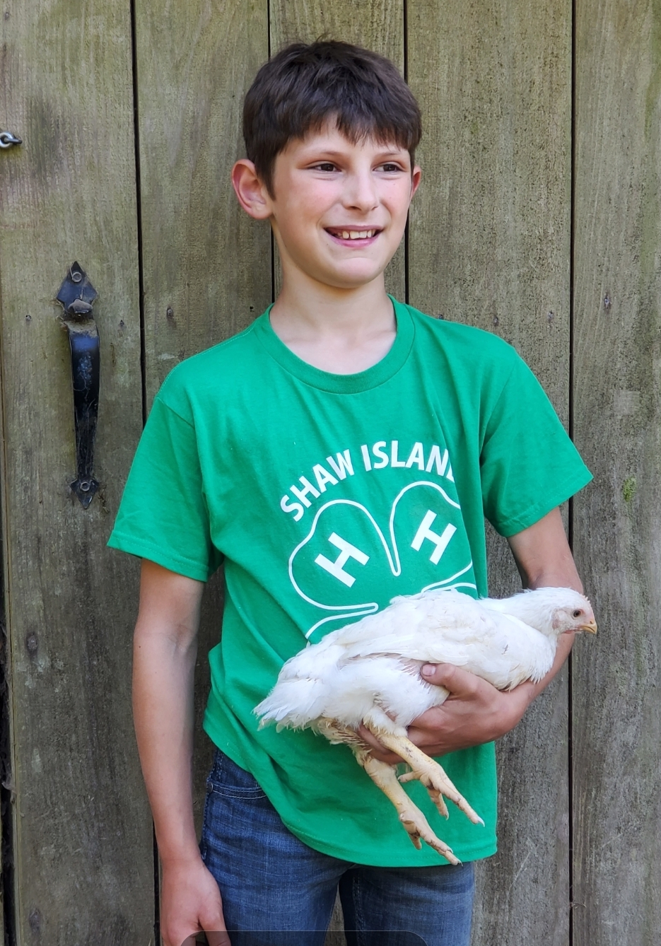 A young boy wearing a green 4-H t-shirt holds a white broiler chicken in front of a wooden door.