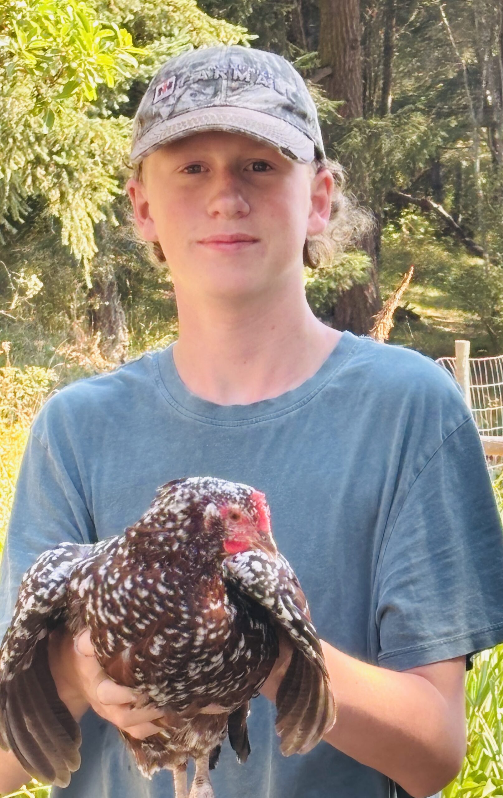 A young man wearing a camo baseball cap and blue t-shirt holds a speckled brown and white chicken outdoors.