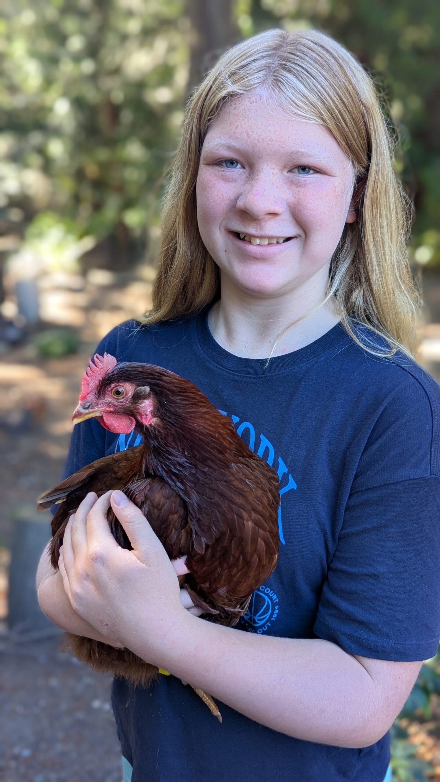 Young 4-H member holding a reddish-brown hen outdoors in a wooded setting.