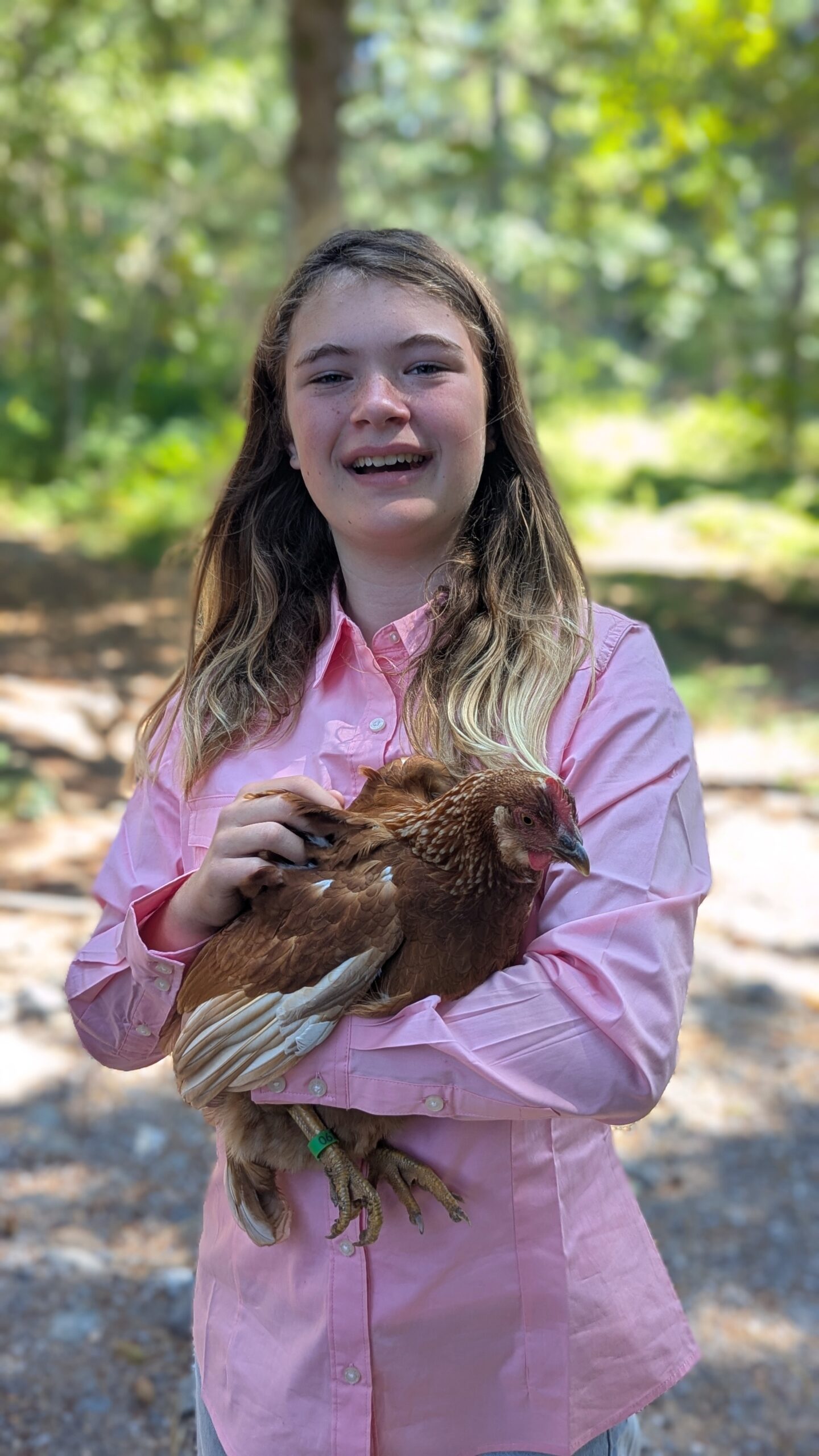 4-H member in a pink shirt holding a brown hen while standing outdoors among trees.
