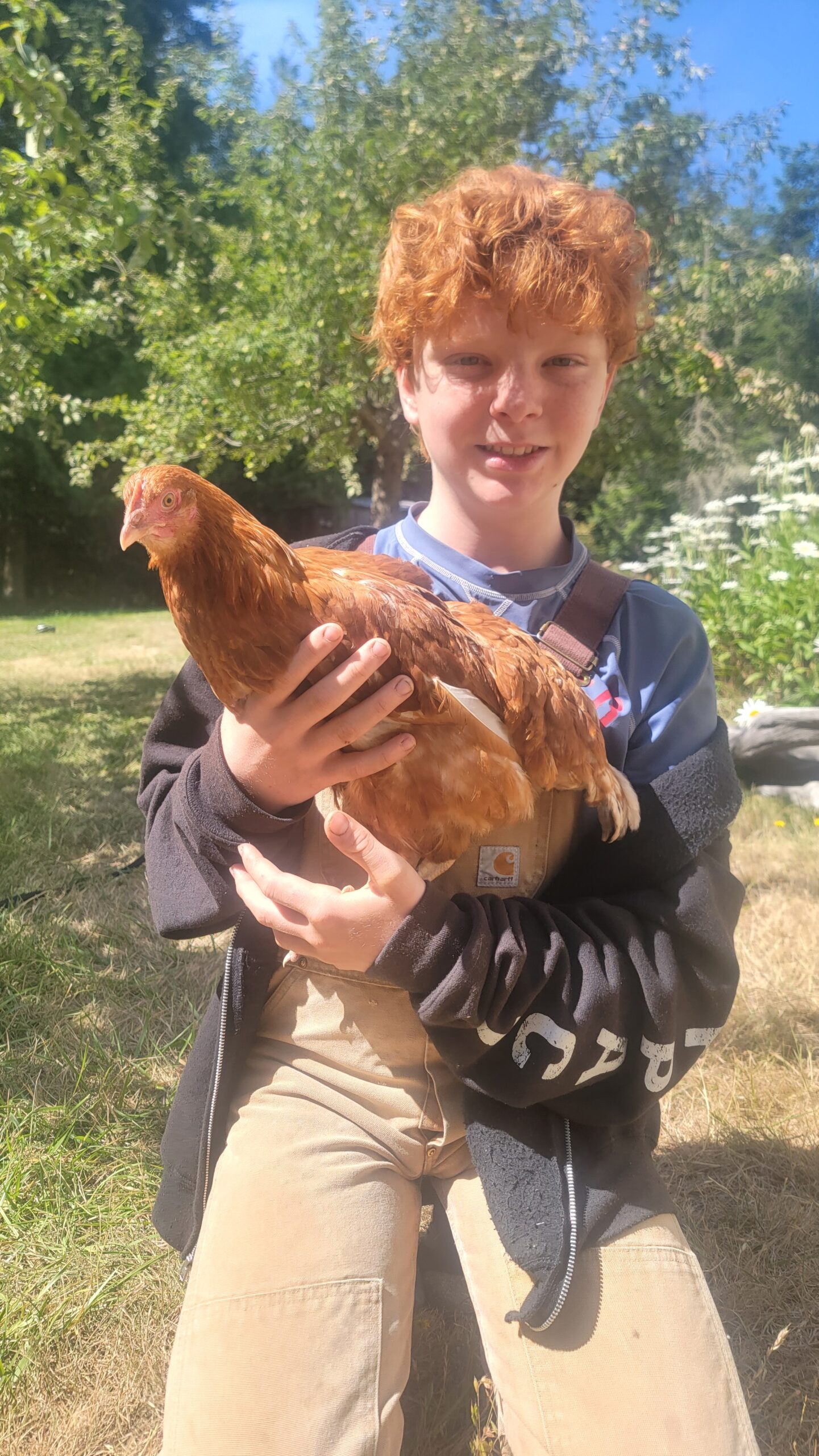A smiling young person with curly red hair holding a light brown chicken outdoors while wearing tan overalls and a black jacket.