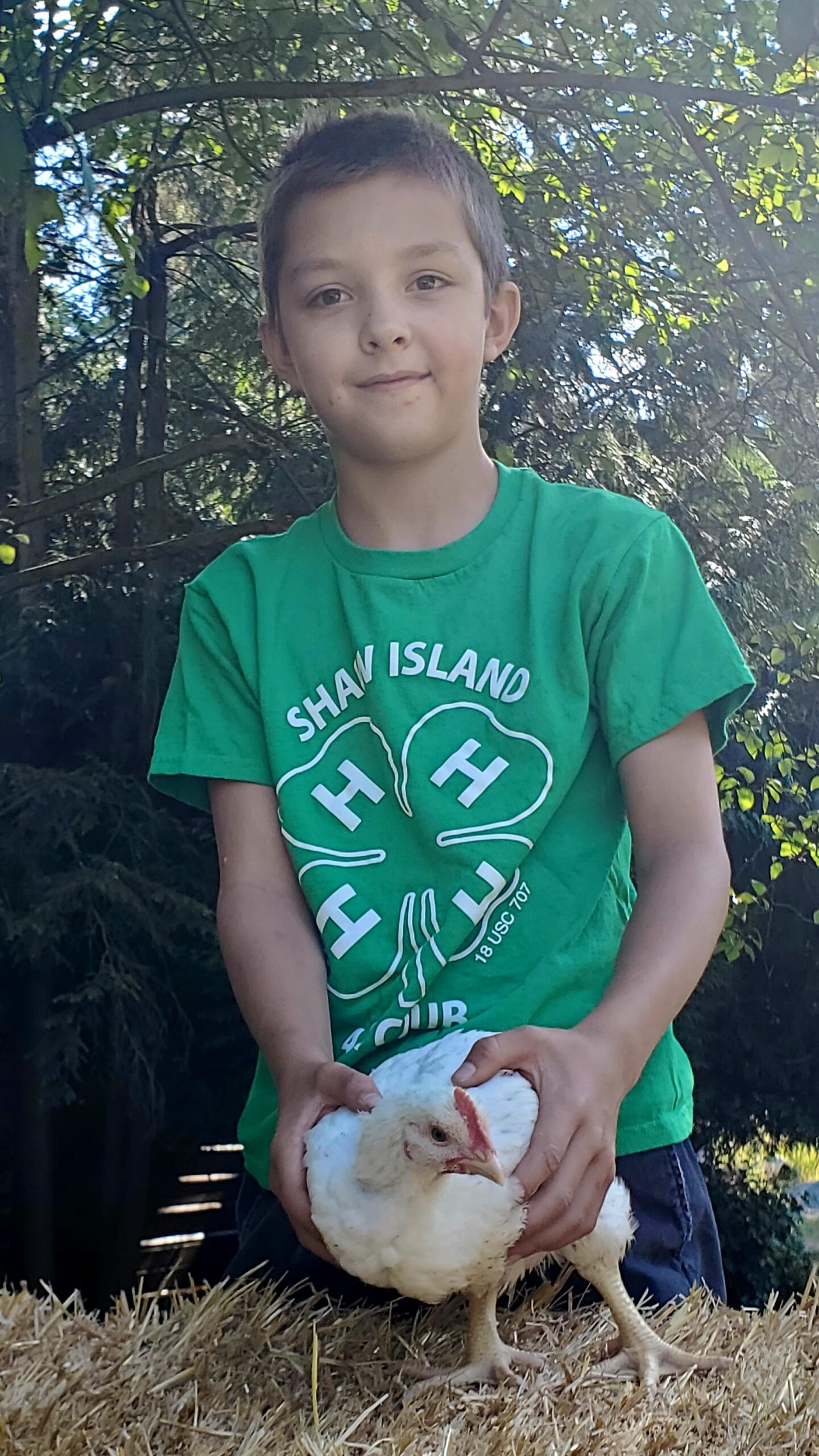 Person wearing a green 4-H Club shirt holding a white chicken outdoors with trees in the background.