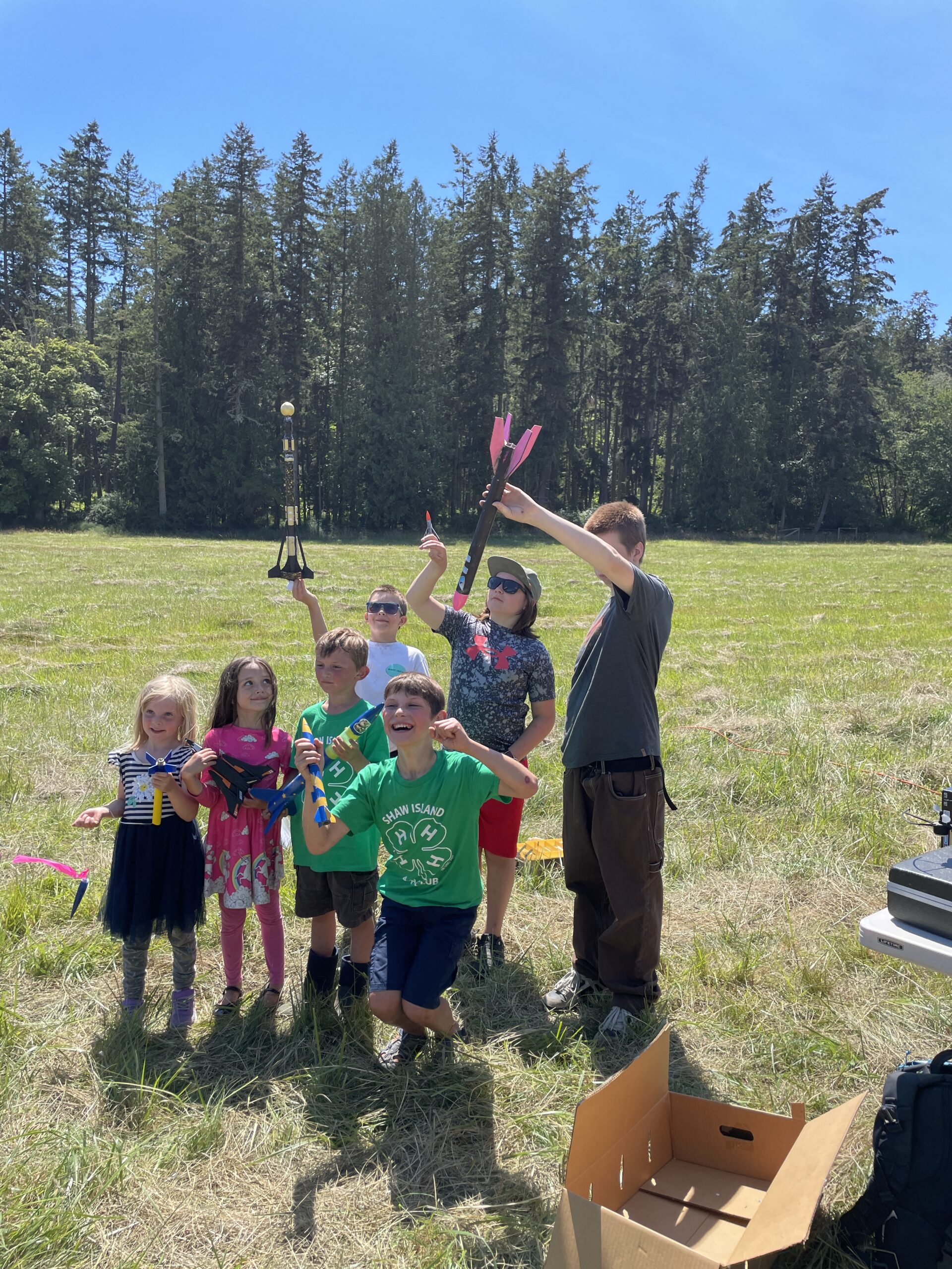 A group of kids standing outdoors in a grassy field on a sunny day, holding up small model rockets.