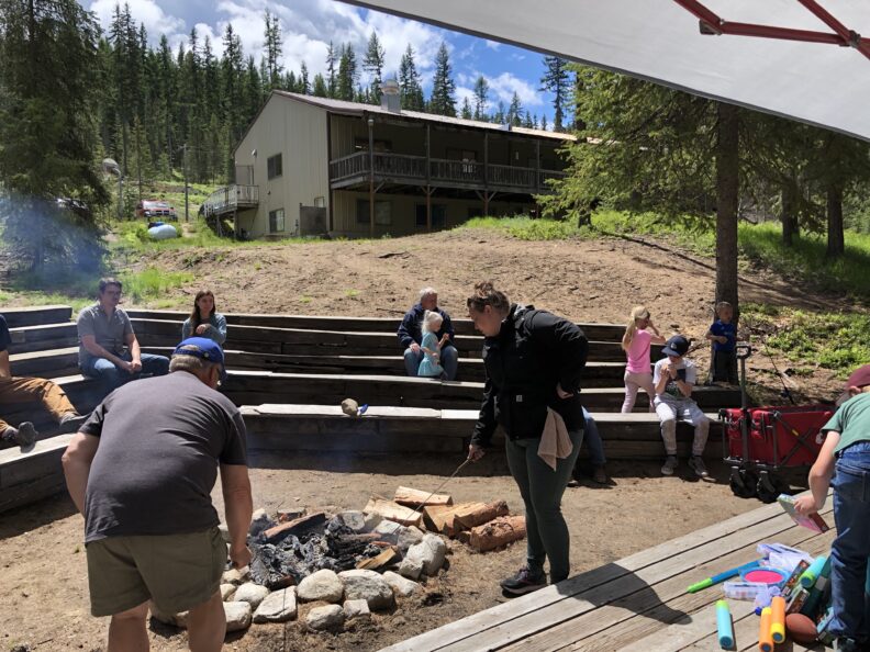 2 adults tend a campfire with kids and adults on benches around the campfire and a building in the background at Camp Princess Pine.