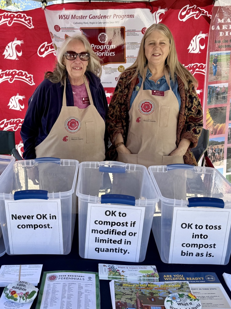 Smiling Master Gardeners Jackie and Kim ready to answer questions at a market booth with a display of what is OK to compost and not OK to compost.
