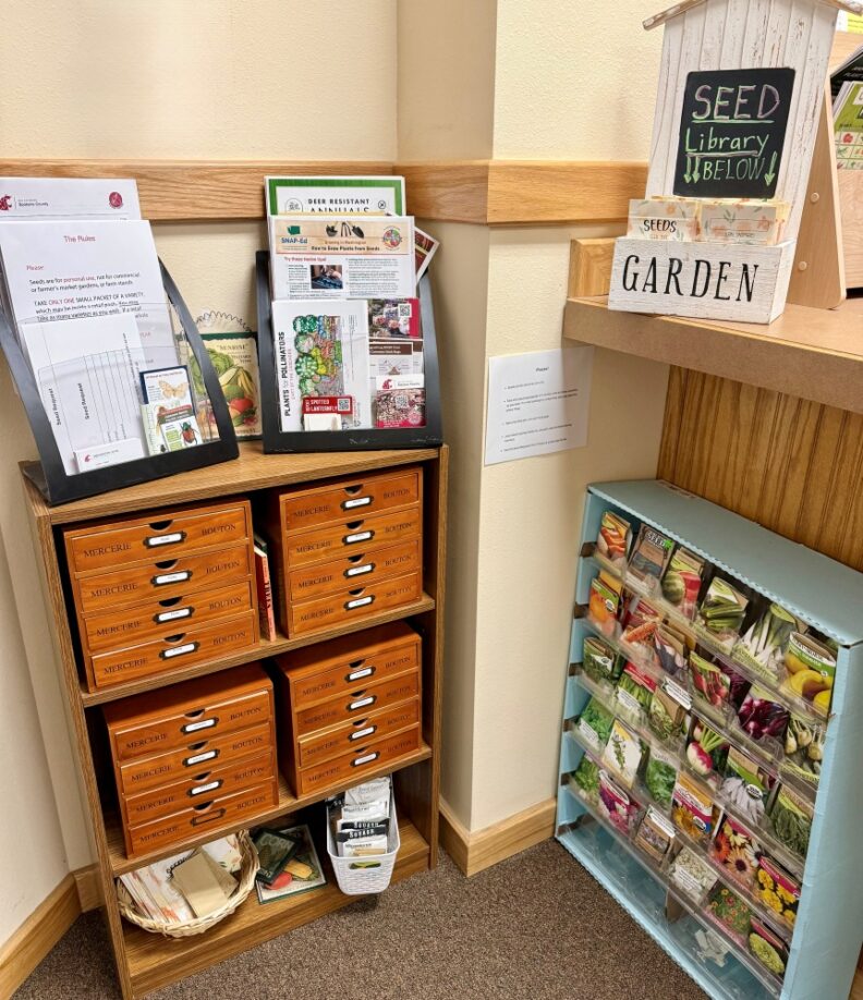 Seed Library in the corner of the lobby of the Colville WSU Extension office, picture shows cabinets of drawers with handouts perched on top and a rack of many different packets of seeds